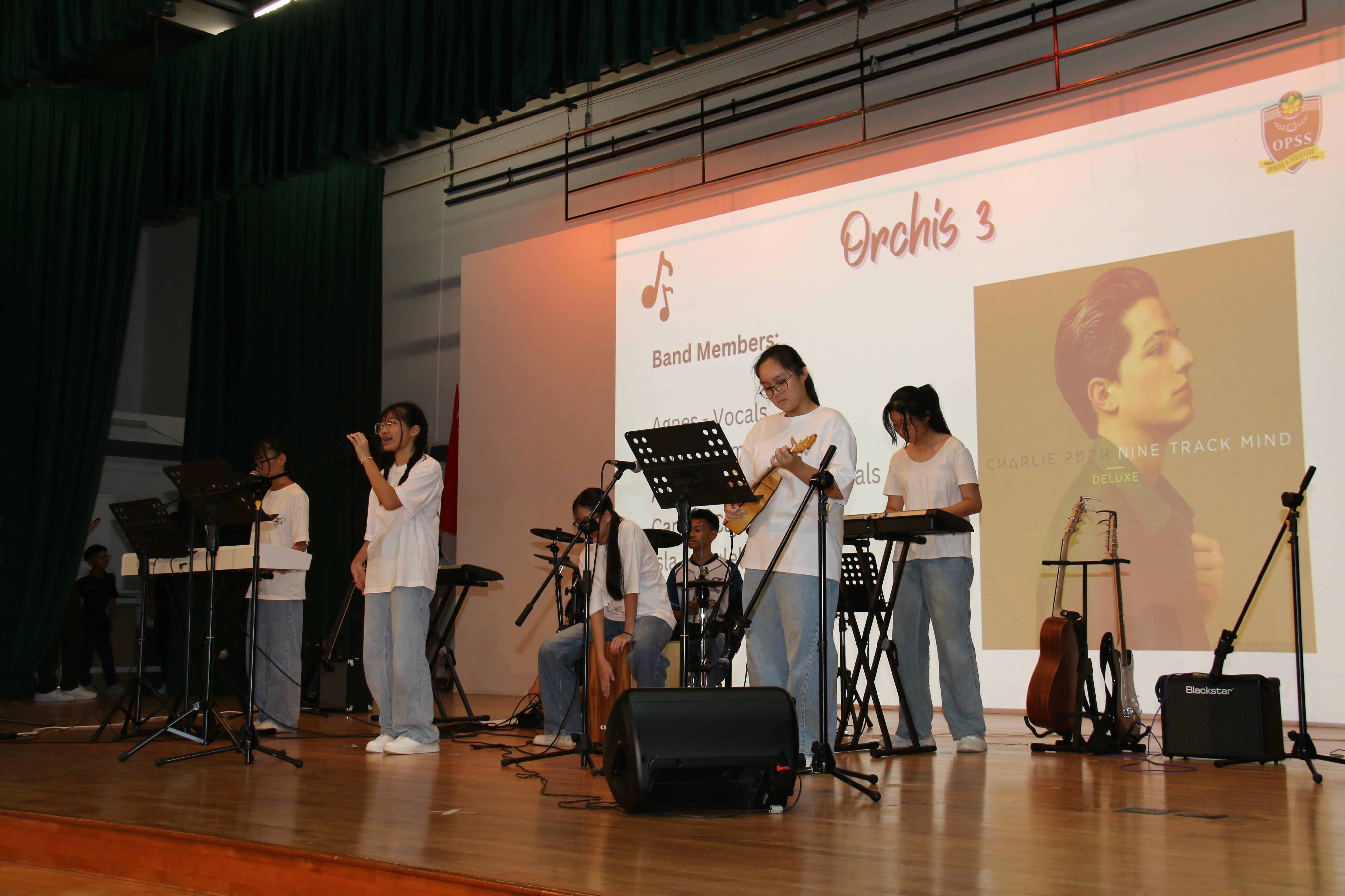 Secondary 2 students performing on school stage during a Battle of the Bands competition. The student band, named "Orchis 3," includes two vocalists, a ukulele player, a keyboardist, and a percussionist on a cajon. A large projector screen behind them displays the band's name and a Charlie Puth album cover.